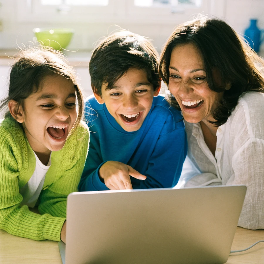 A mother and her two children laughing together at a laptop at the kitchen table