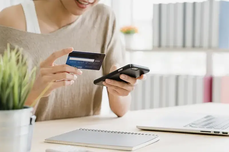 Close-up of a person holding a credit card and smartphone, making an online payment, with a laptop and notepad on a desk, symbolizing secure online transactions or digital learning resource purchases