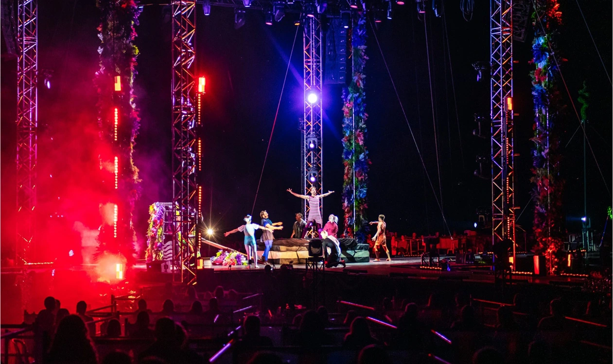 A nighttime stage with colourfully-dressed performances and lights, smoke, and attendees near the stage. 
