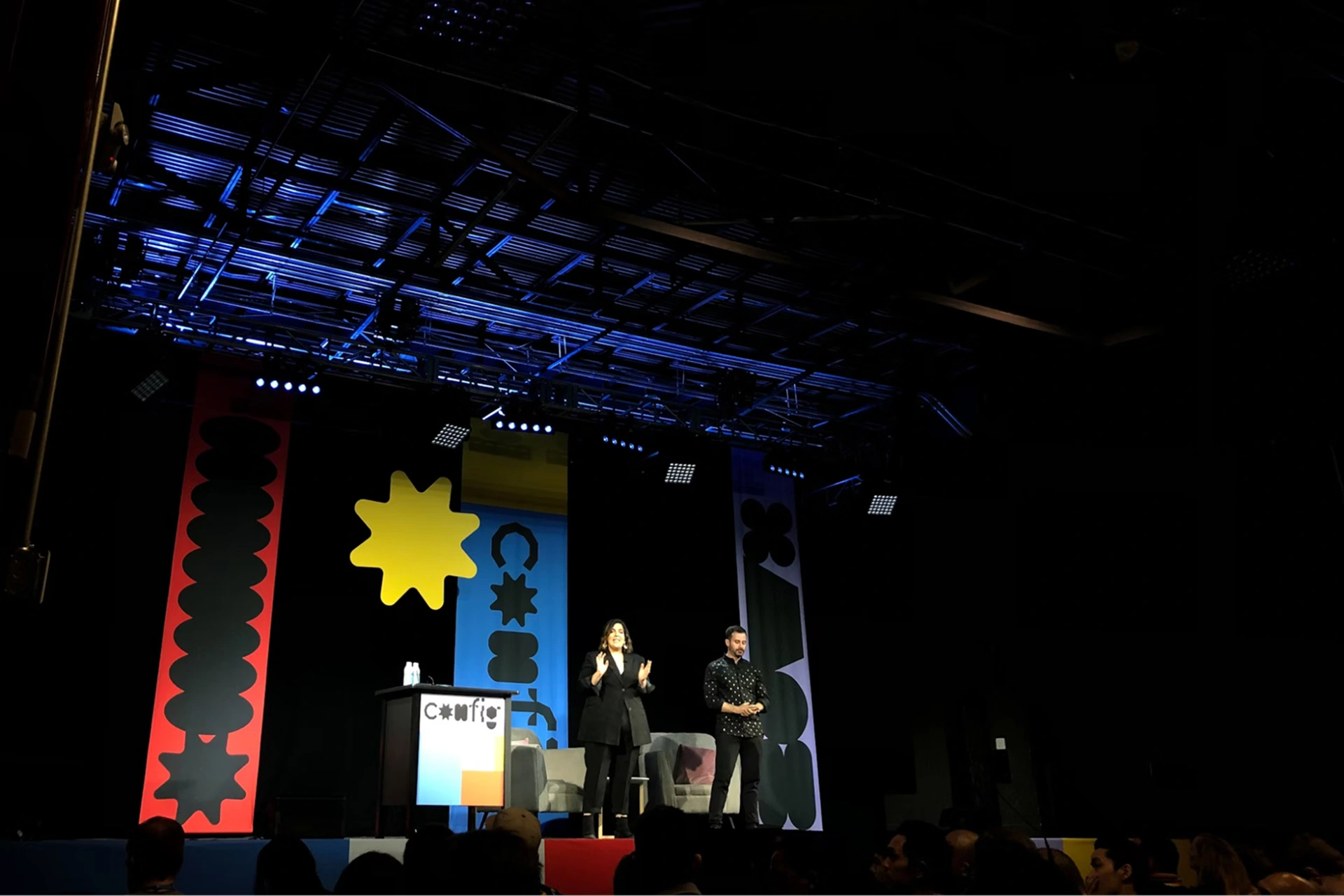 Two presenters stand on a stage in a darkened conference room, with posters behind them. 