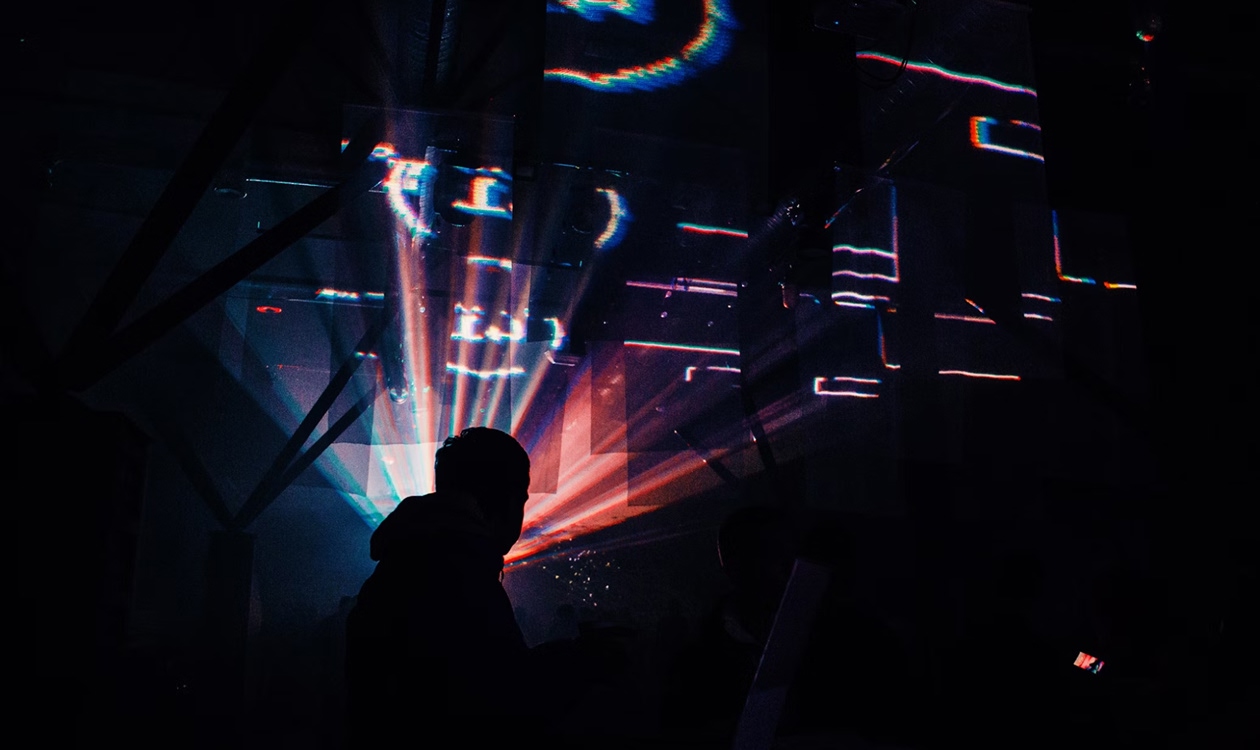 A male silhouette stands before a colourful holographic projection overhead amidst a dark room. 