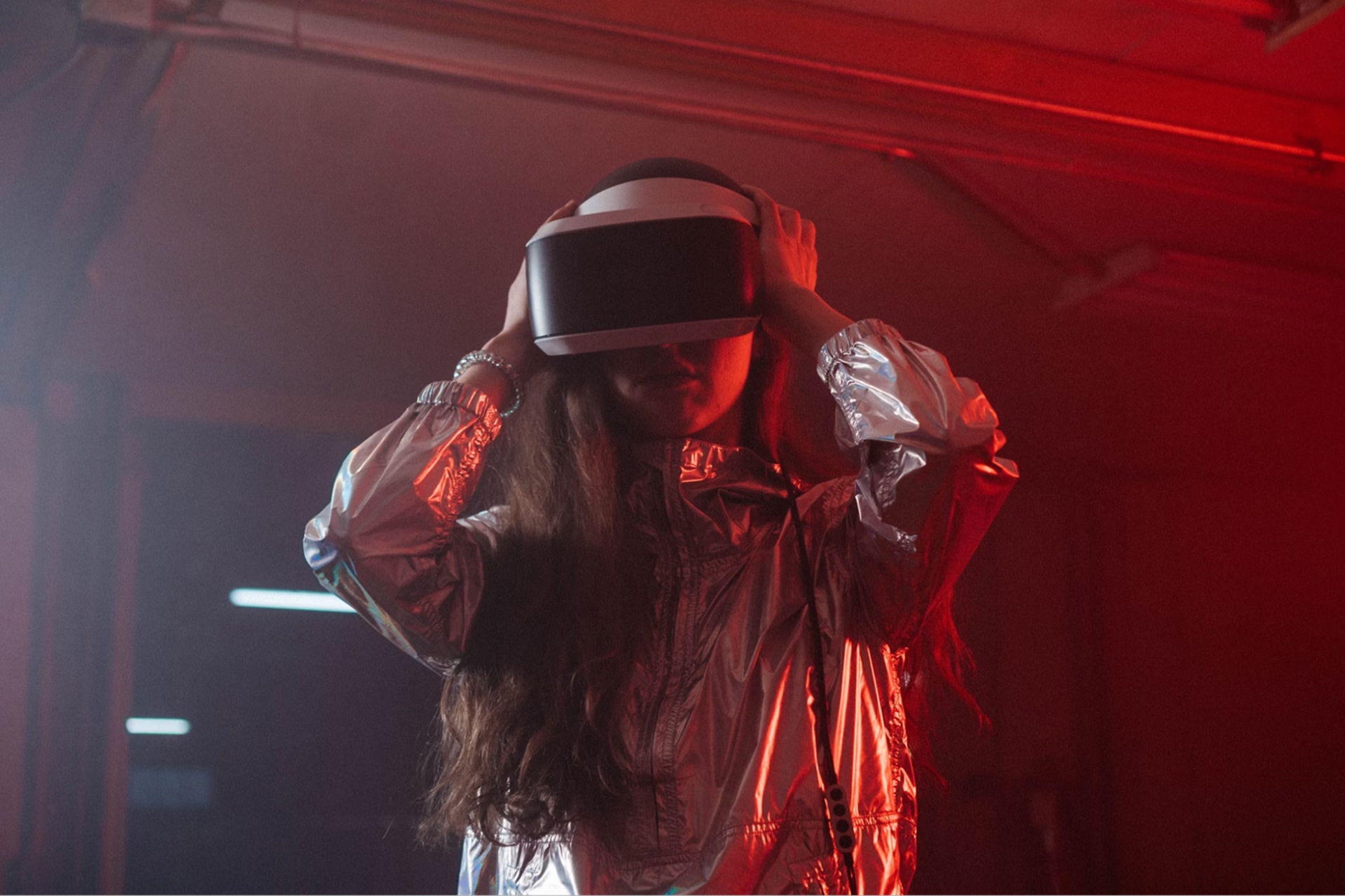 A woman wearing a silver jacket and a VR headset on her head in a red-lit room. 