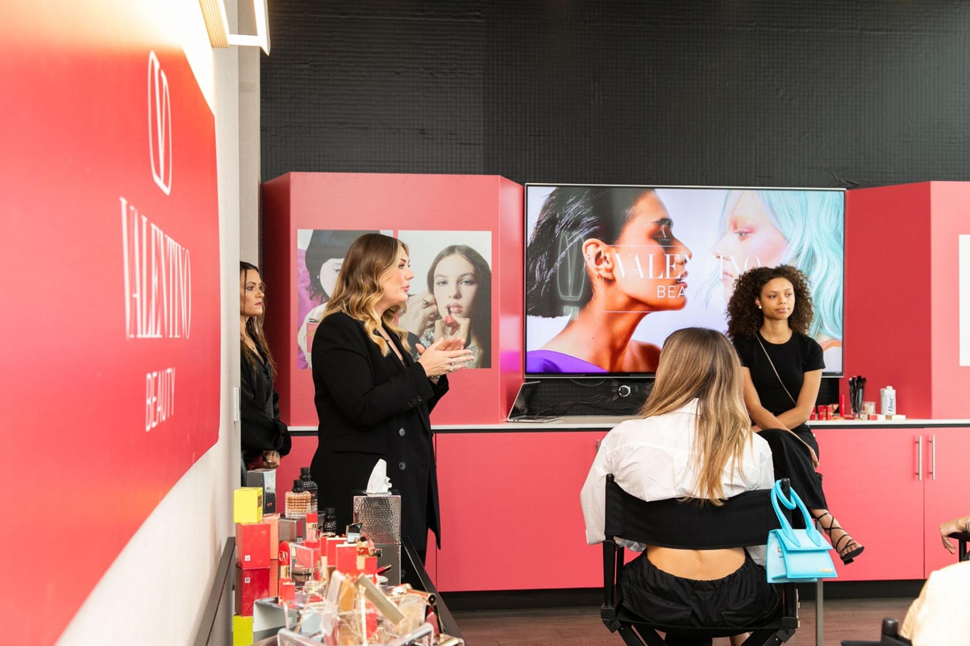 Two women stand at a makeup display booth, one demos to two women seated at chairs. 
