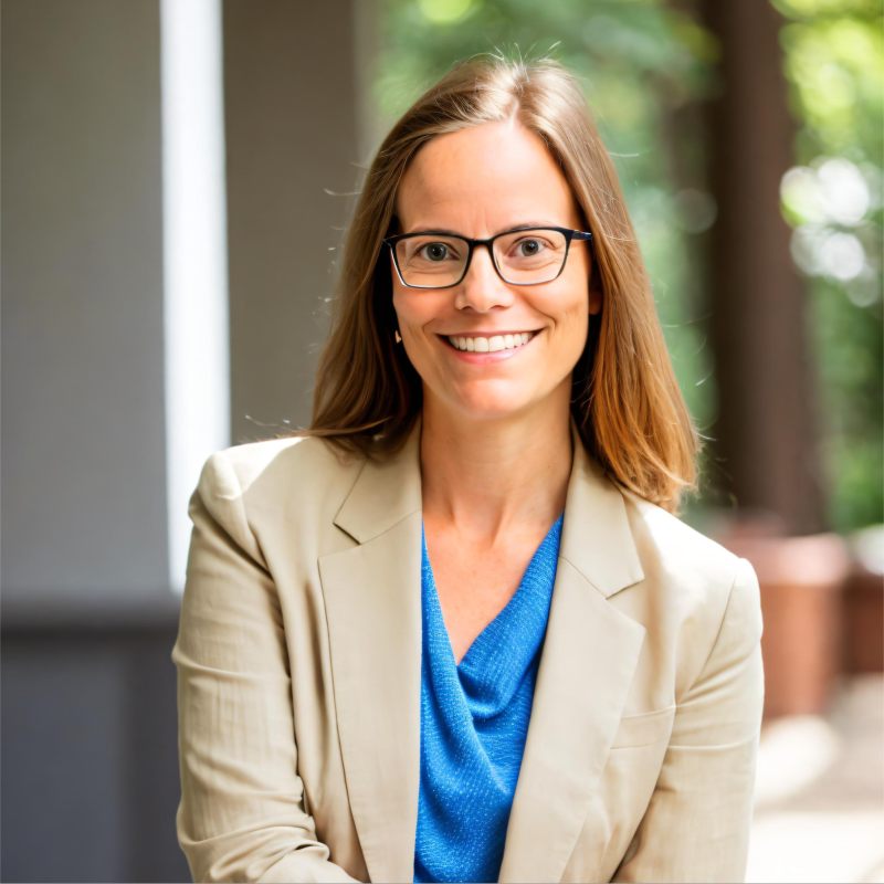 Smiling woman with light brown hair and glasses wearing a beige blazer over a blue top, standing outdoors with blurred greenery and building in the background.