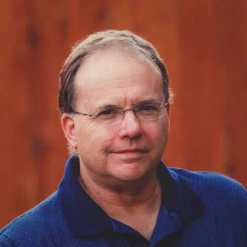 Middle-aged man with glasses and thinning hair, wearing a dark blue collared shirt, standing against a blurred wooden background.