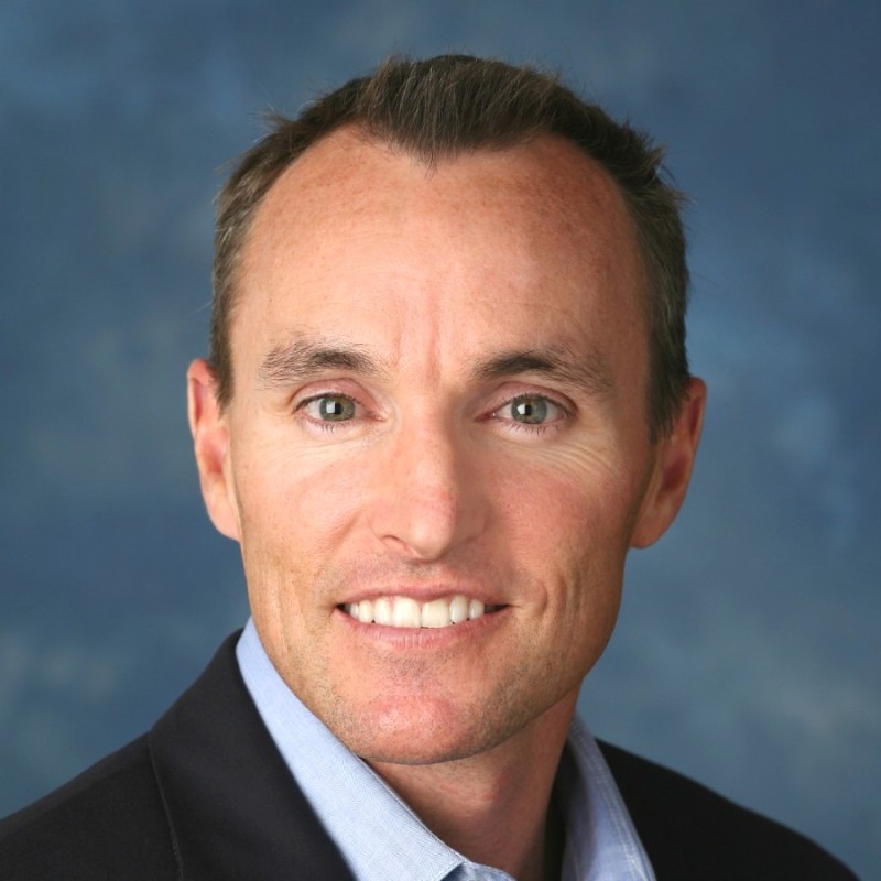 A headshot of a middle-aged man with light skin and short brown hair, smiling and wearing a dark blazer over a light blue collared shirt, with a blurred blue-grey background.