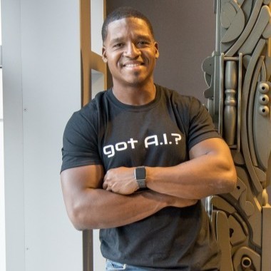 Smiling man with crossed arms wearing a black T-shirt that says 'got A.I.?' and a smartwatch, standing indoors near a decorative panel.