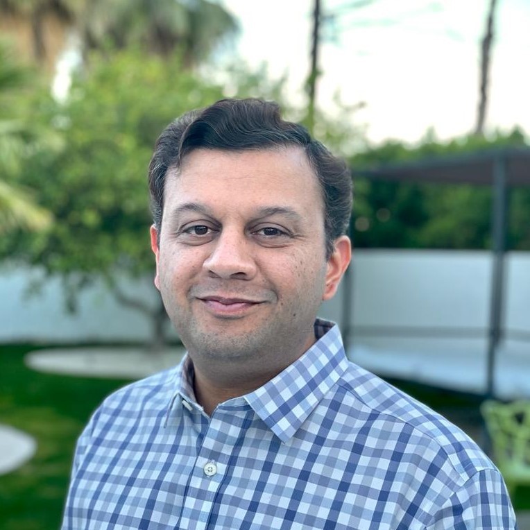 Man with short dark hair wearing a light blue and white checkered shirt standing outdoors with greenery and trees in the blurred background.