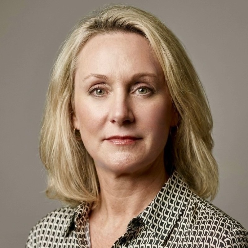 Portrait of a middle-aged woman with shoulder-length blonde hair, light makeup, wearing a patterned collared shirt, looking directly at the camera with a neutral expression against a plain background.