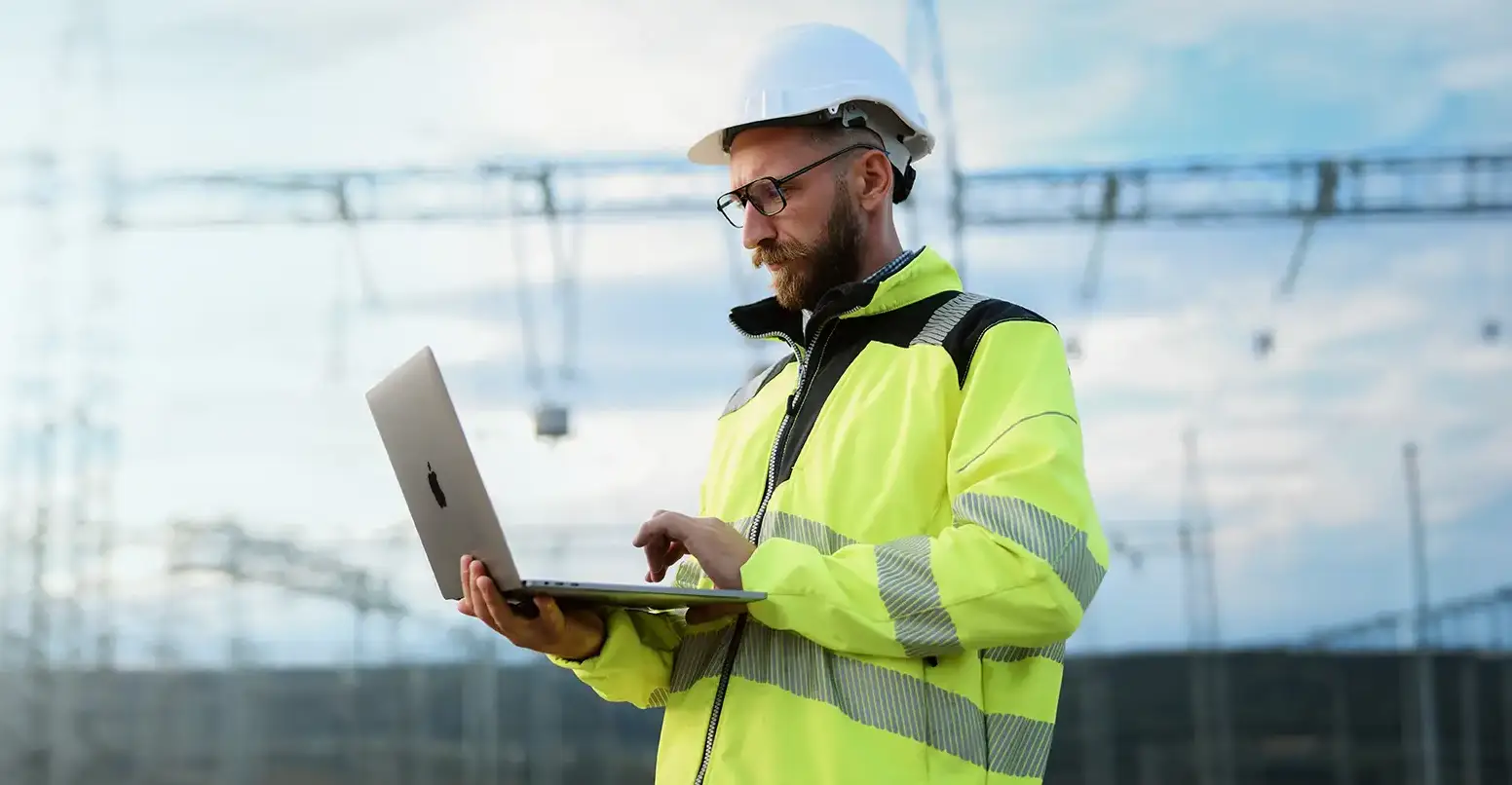 Construction worker wearing a white hard hat and yellow reflective jacket using a laptop outdoors at a construction site.
