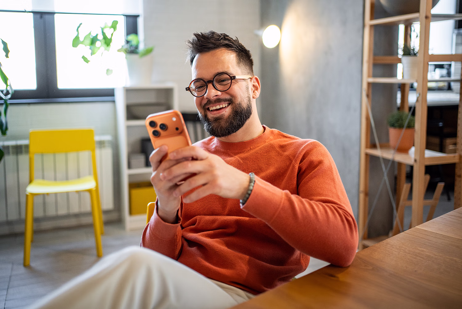 Smiling man with glasses and beard sitting indoors looking at smartphone.