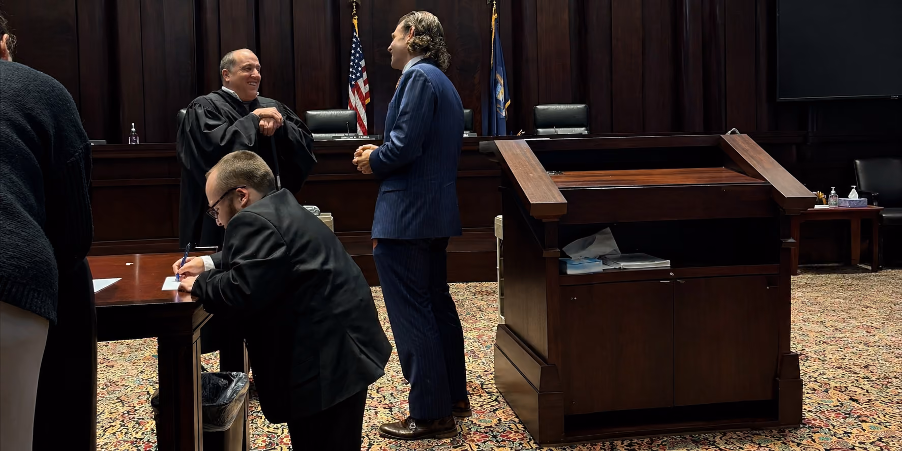 Man in judge robe talks with two men in suits inside a courtroom with flags and wood paneling.