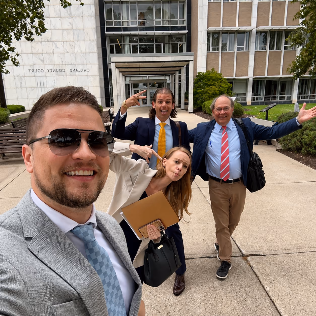 Four people in business attire smiling and posing joyfully outside Oakland County Court building.