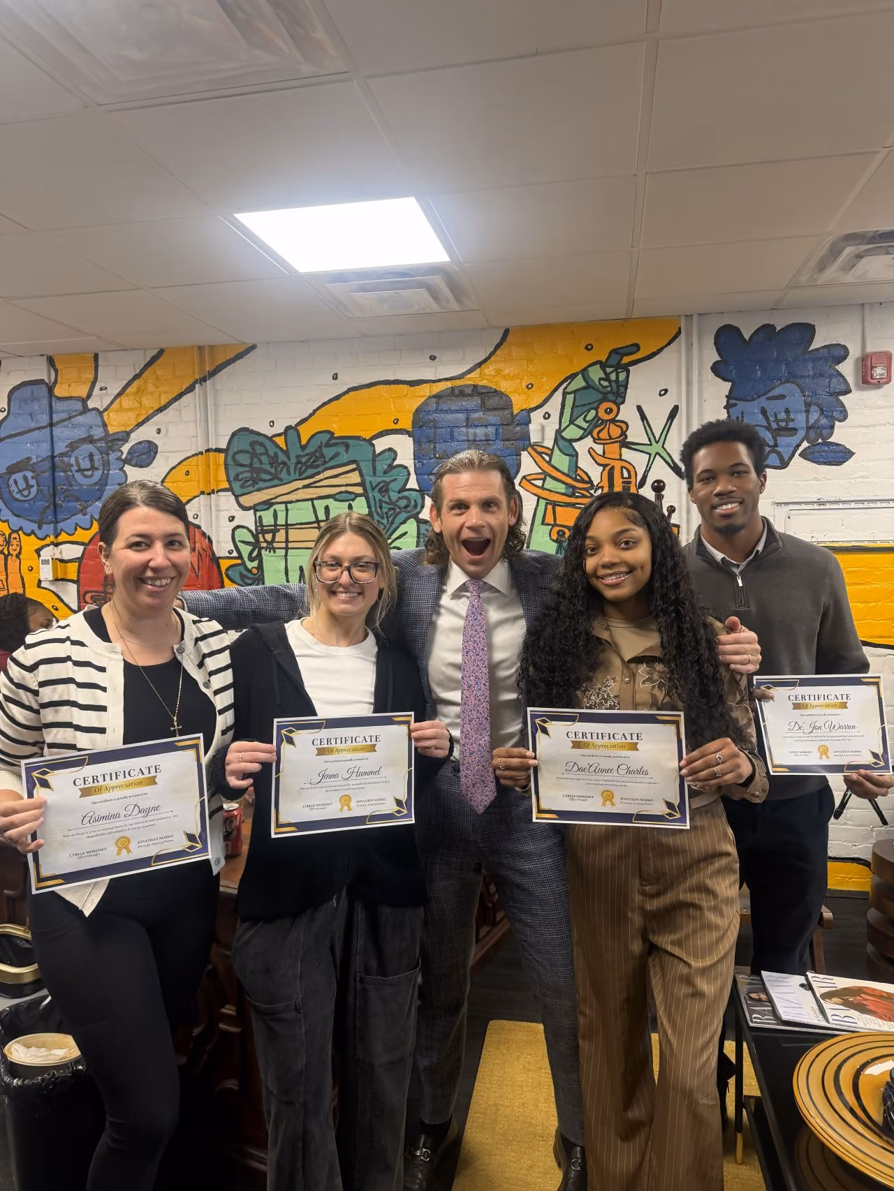 Five people standing indoors holding certificates of appreciation, smiling in front of colorful mural wall art.