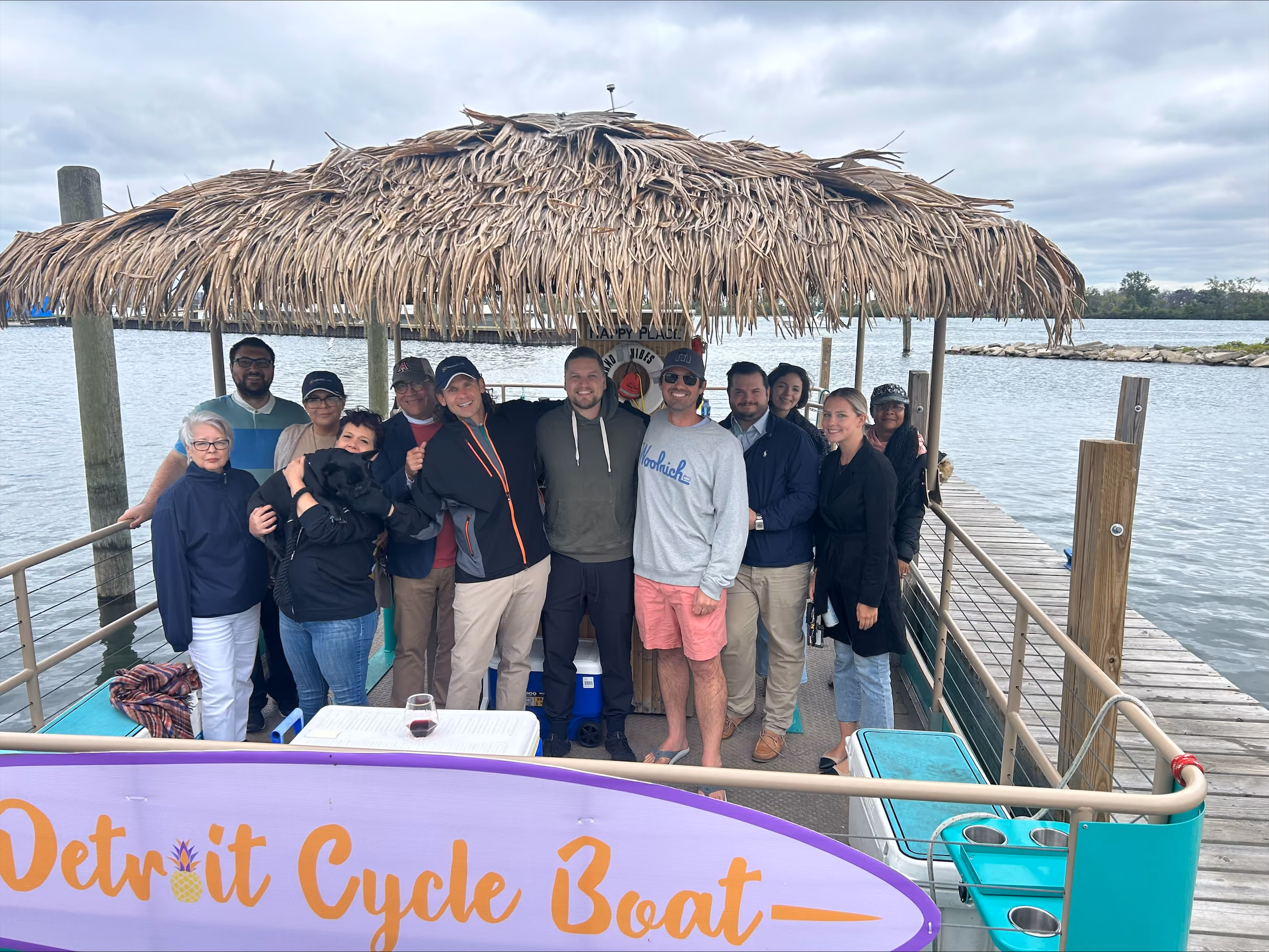 Group of people standing on a Detroit Cycle Boat with a thatched roof by a lakeside dock.