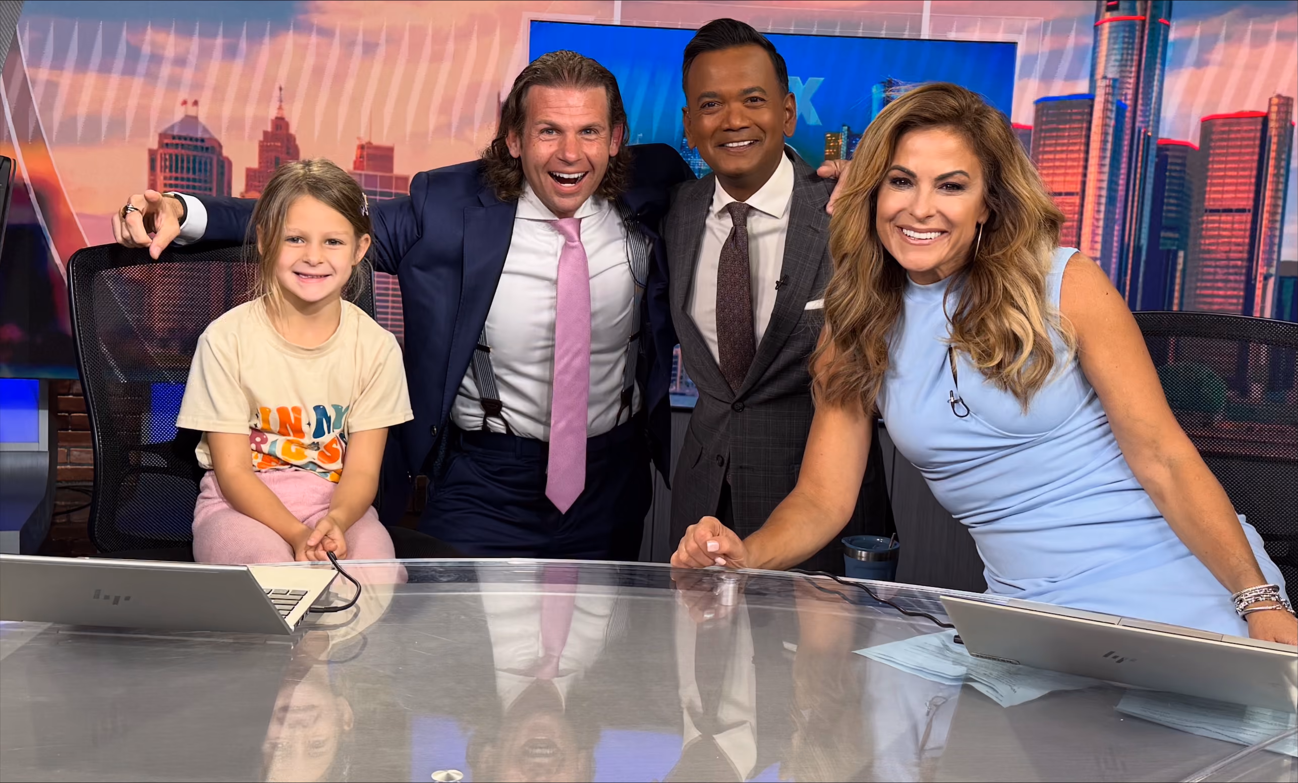 Four people, including a smiling young girl, two men in suits, and a woman in a blue dress, pose at a news desk.