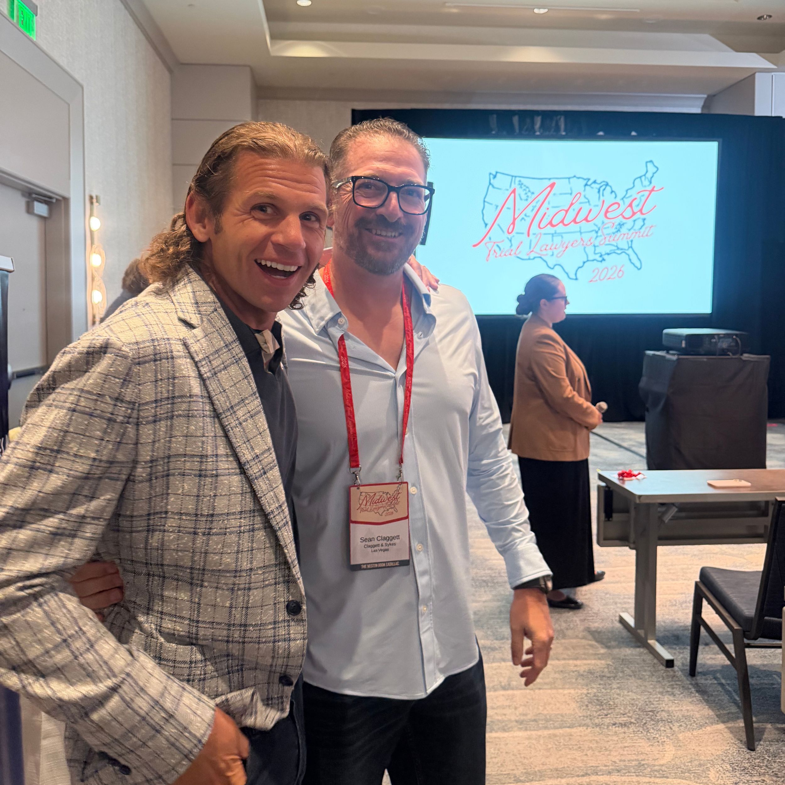 Dos hombres sonríen para una foto frente a una pantalla grande que muestra "Midwest Fall Savings 2020" con un mapa de los Estados Unidos.