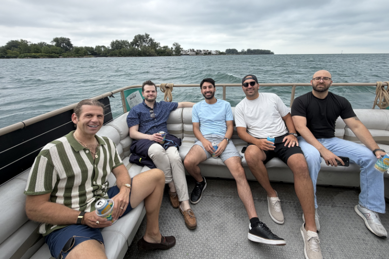 Five men sitting on a boat bench holding drinks with water and trees in the background under cloudy sky.
