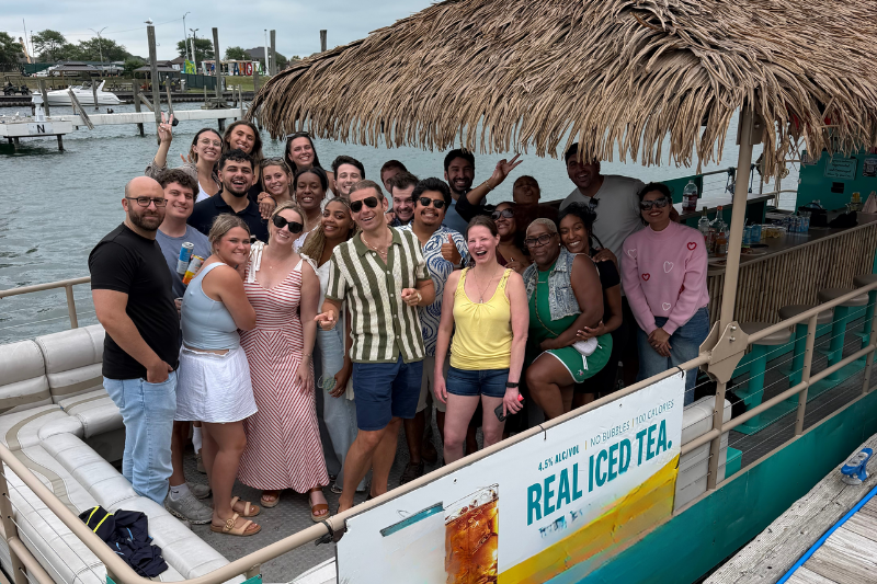 Group of diverse people smiling and posing together on a boat with a thatched roof by the water.