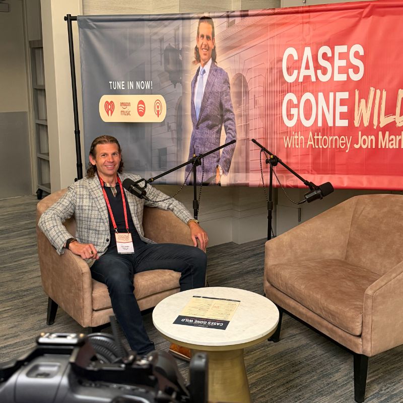 Jon Marko with lanyard seated for podcast recording with banner reading Cases Gone Wild and microphones