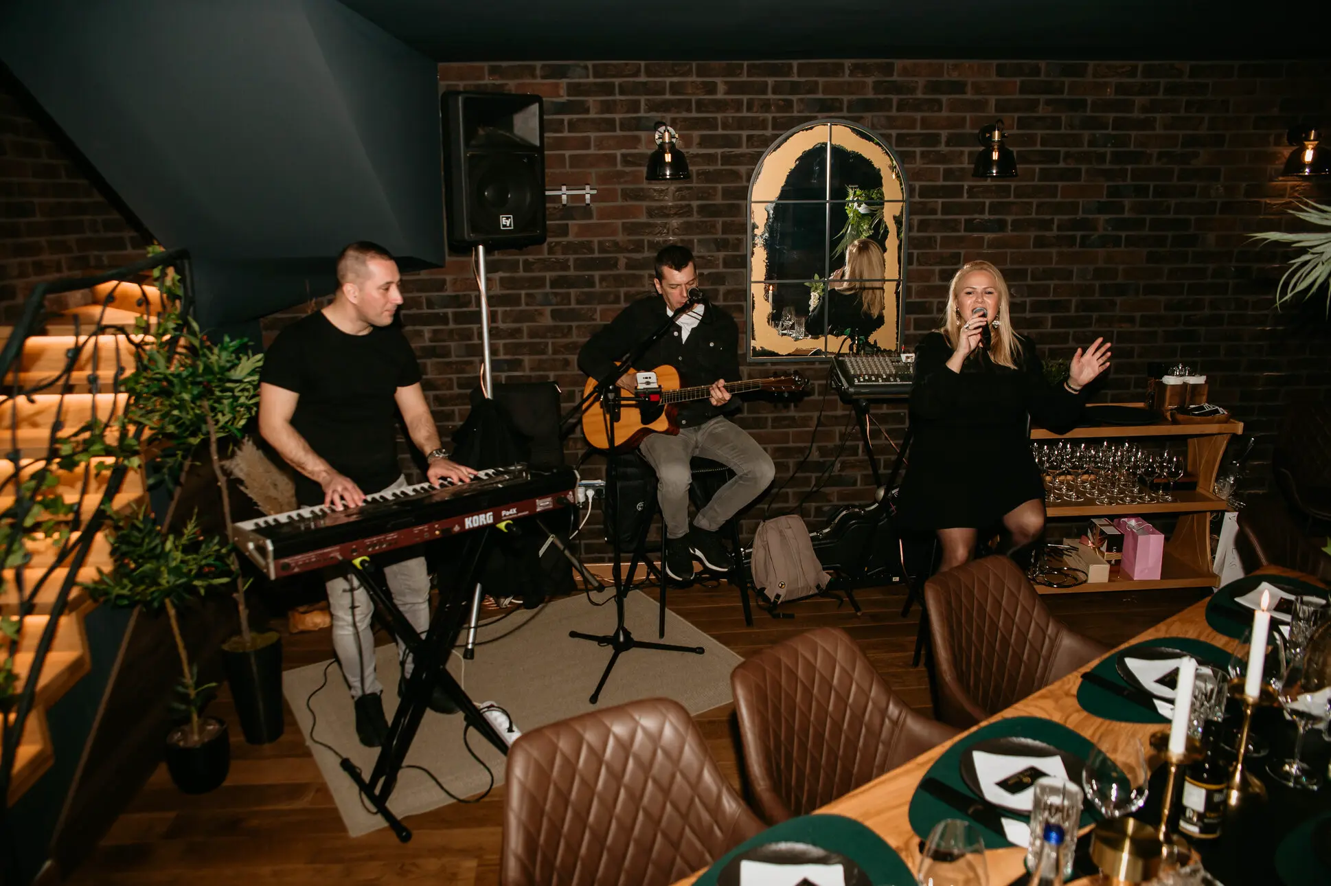Tres miembros de la banda en un espacio interior con una pared de ladrillos, un hombre toca el teclado, otro toca la guitarra acústica y una mujer canta con un micrófono en la mano.