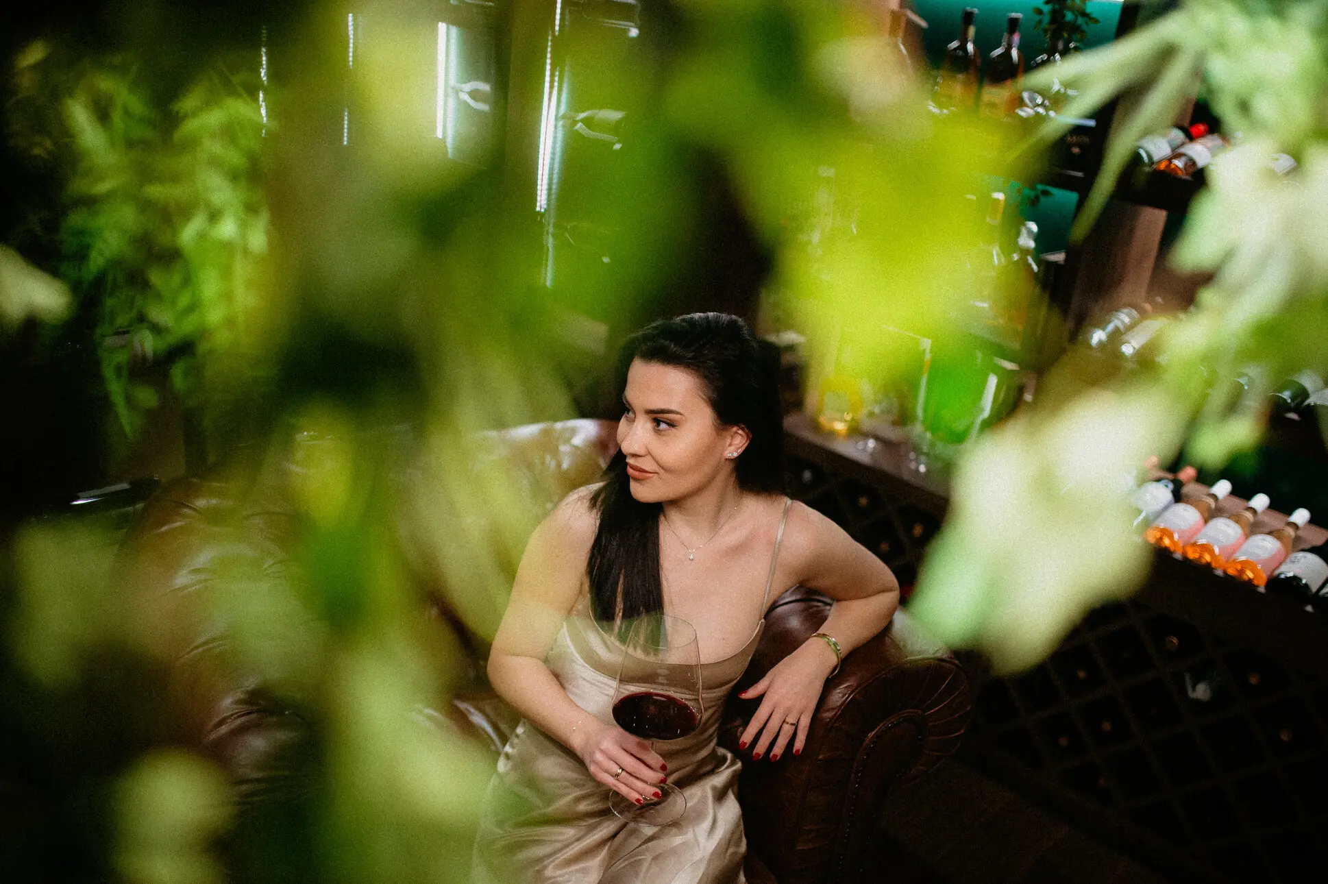 Una mujer de pelo largo y negro con un vestido plateado se sienta en un sillón de cuero y sostiene una copa de vino tinto en la mano en una bodega bellamente iluminada.