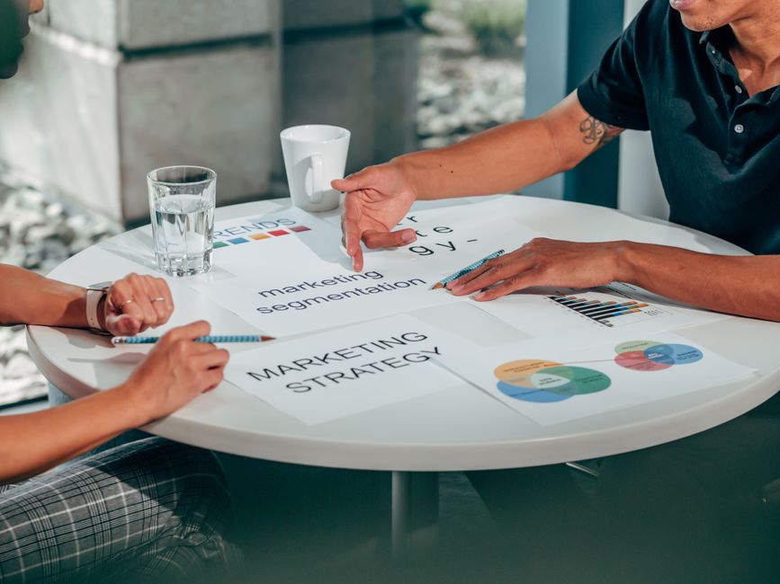 Two colleagues discussing a marketing strategy plan around a table, representing a business making its first marketing hire.
