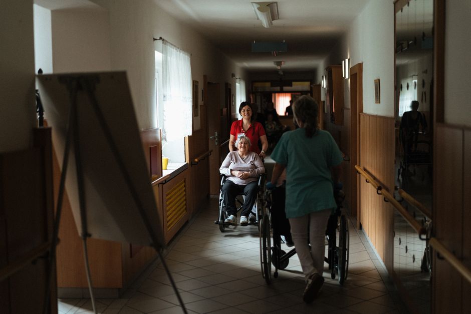 Caregiver assisting an elderly woman in a wheelchair along a retirement home corridor