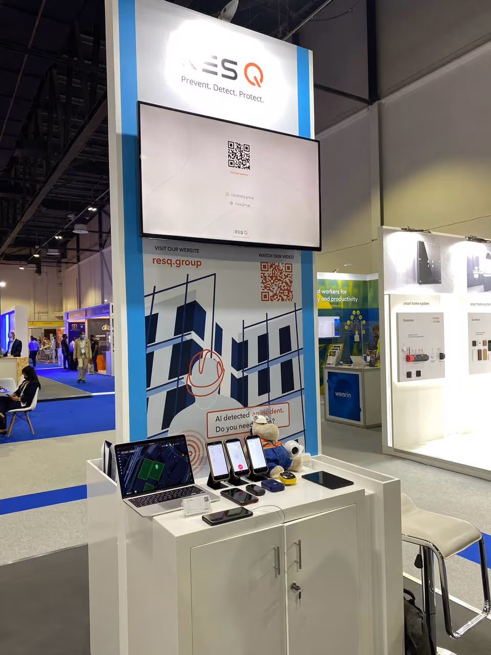 Trade show booth for RESQ featuring a large screen, company logo, QR codes, multiple smartphones, a laptop, and a plush toy on a white counter.