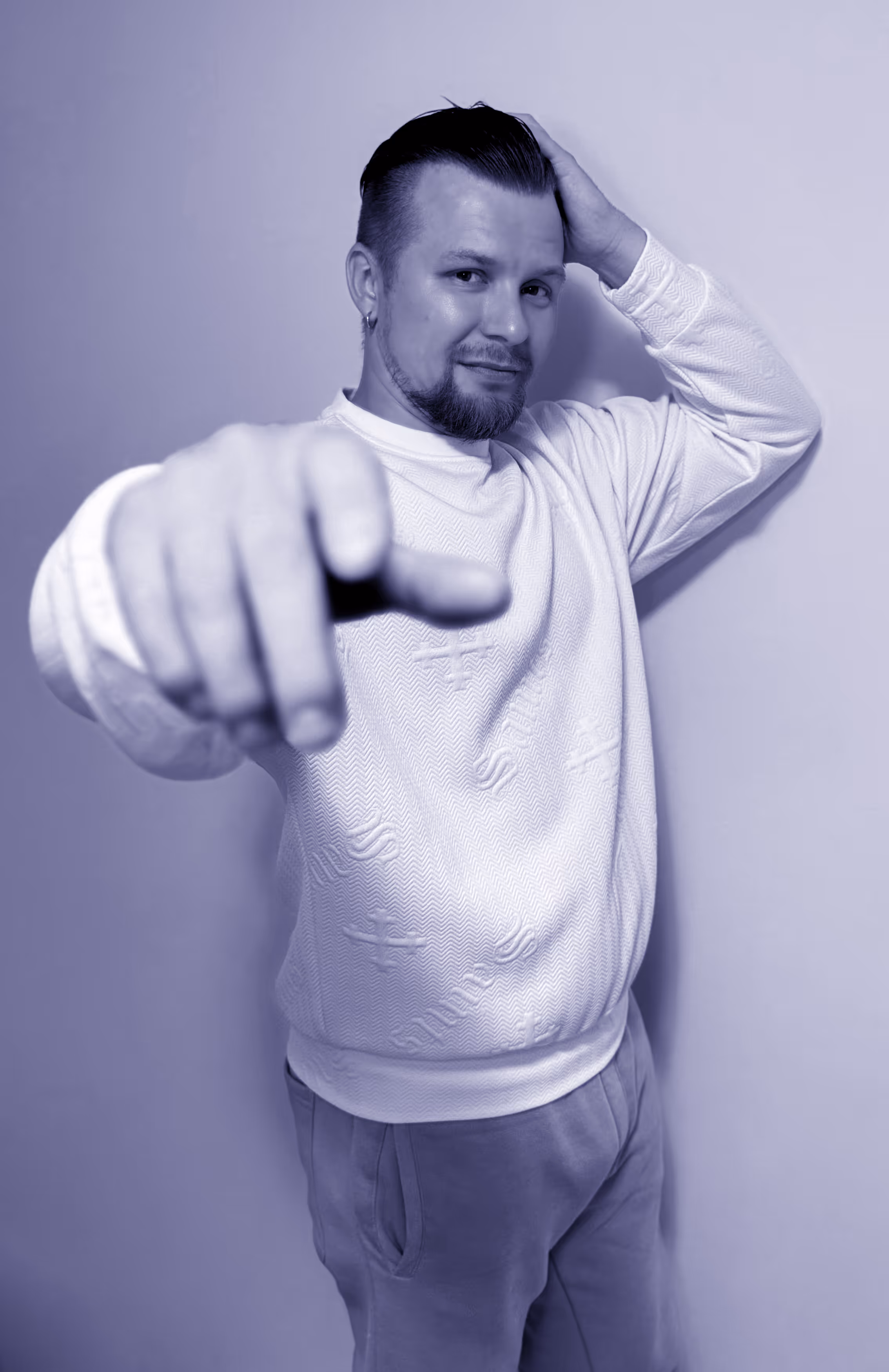 Man with short hair and beard wearing a textured white sweater and casual pants, pointing toward the camera with one hand while touching his head with the other, standing against a plain background