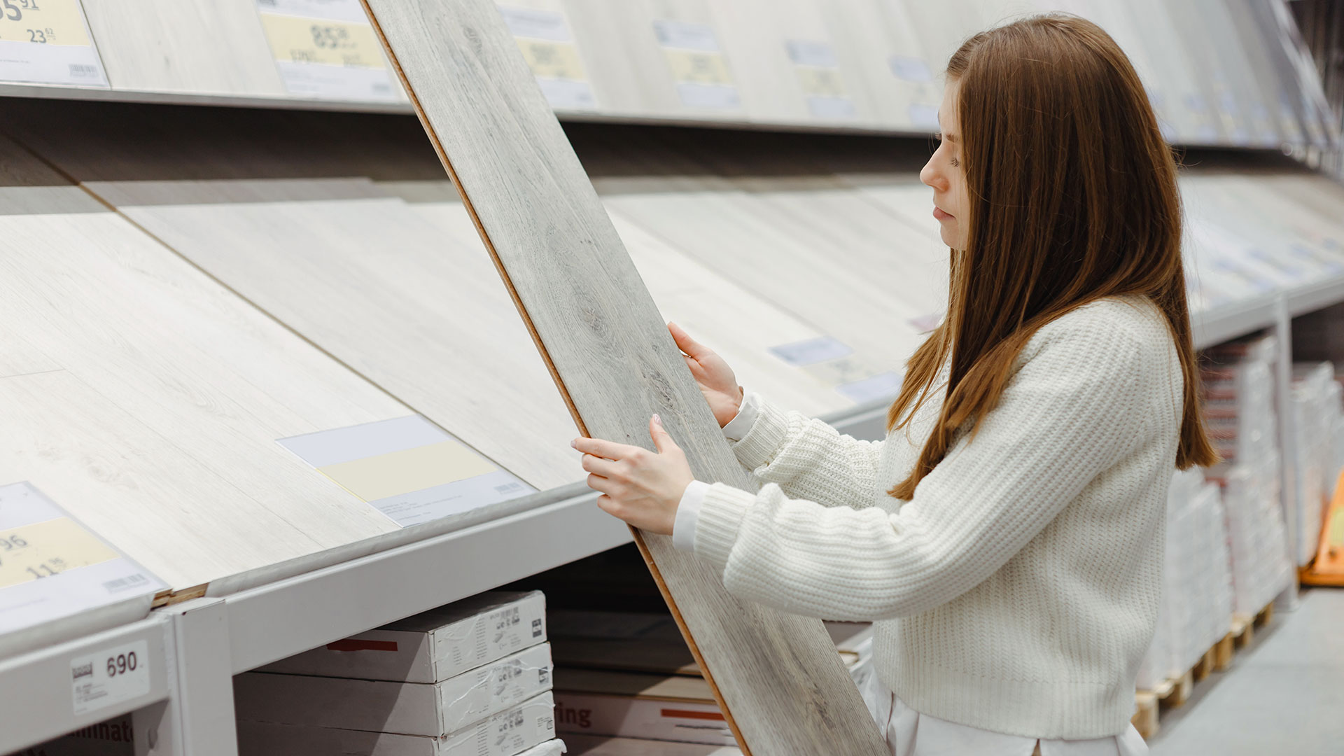 A woman in a showroom holds and examines a sample of gray laminate flooring. She inspects the texture and finish under bright display lighting.