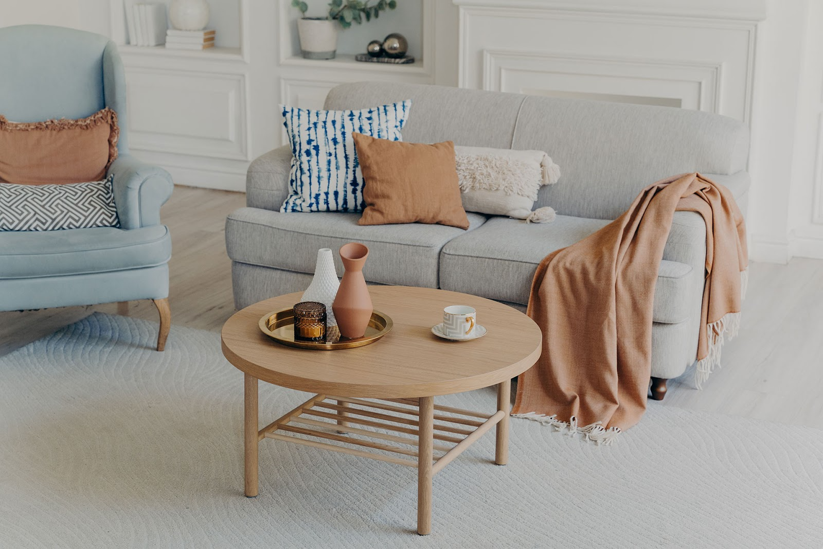 Cosy living room with grey 2-seater couch, blue and terracotta throw cushions and blanket, blue occasional chair, white wall coving, light coloured floor boards.