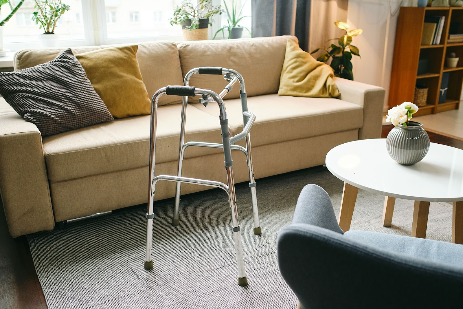 Walking frame in front of a lounge and coffee table, grey rug, timber floors, bookcase and indoor plants on the windowsill.
