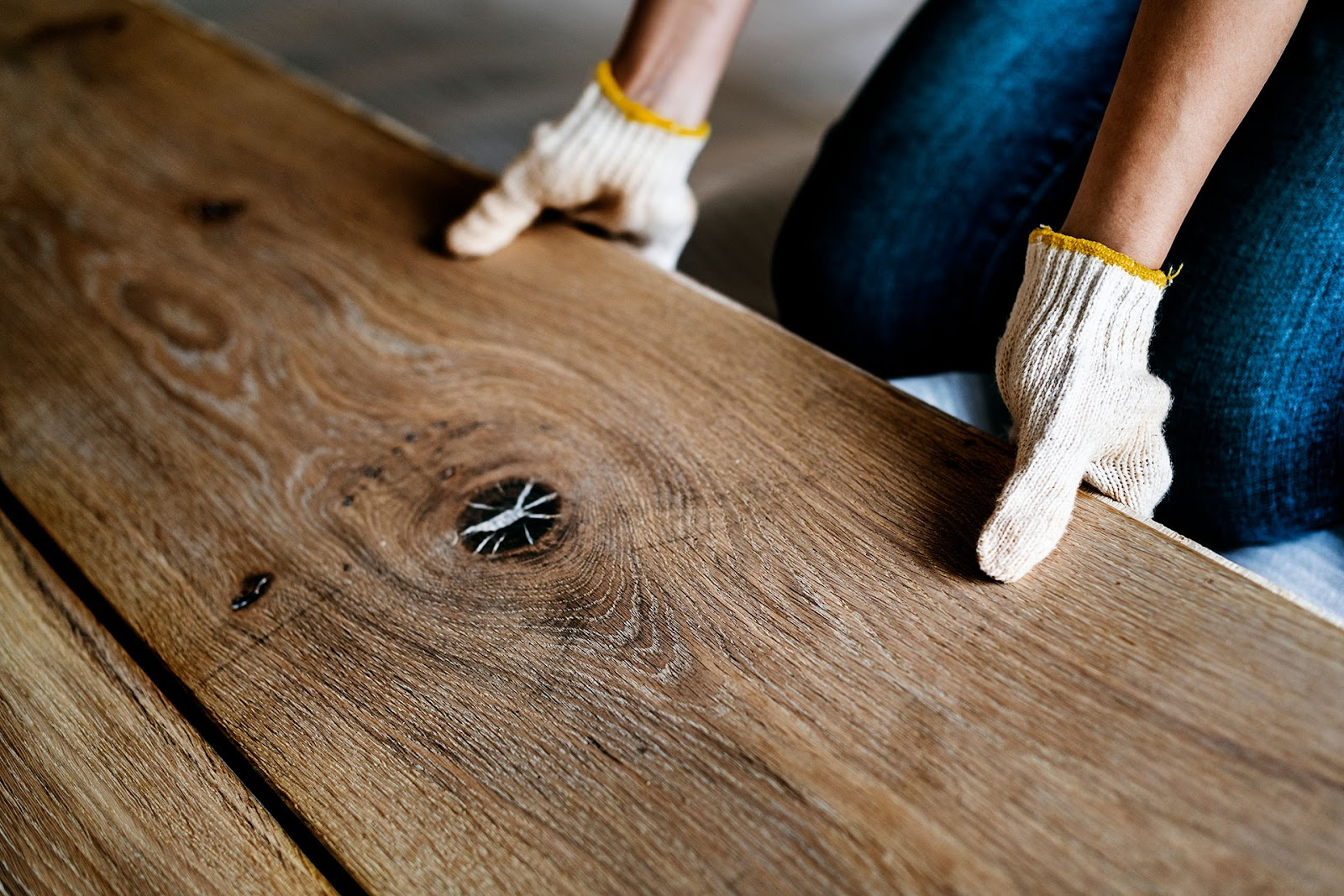 Man in blue jeans and gloves kneeling to install timber hardwood floorboards.