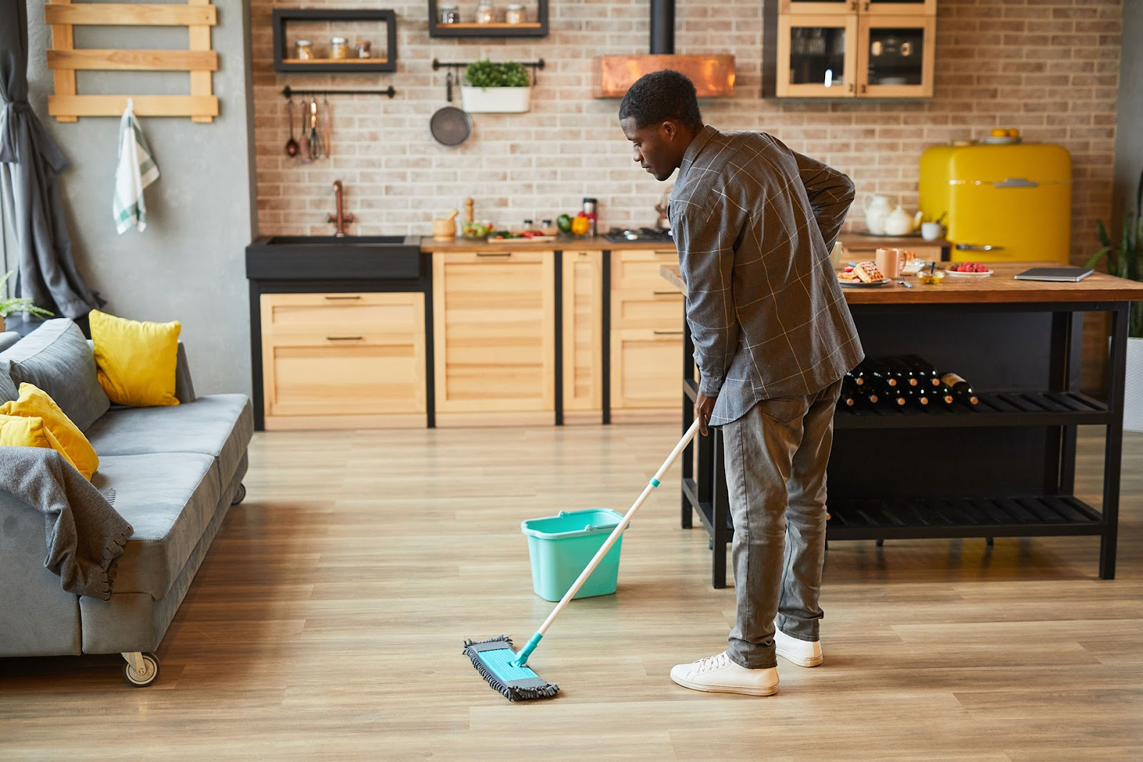 Modern apartment with open plan kitchen, island bench and grey couch. Man mops light timber floor. 
