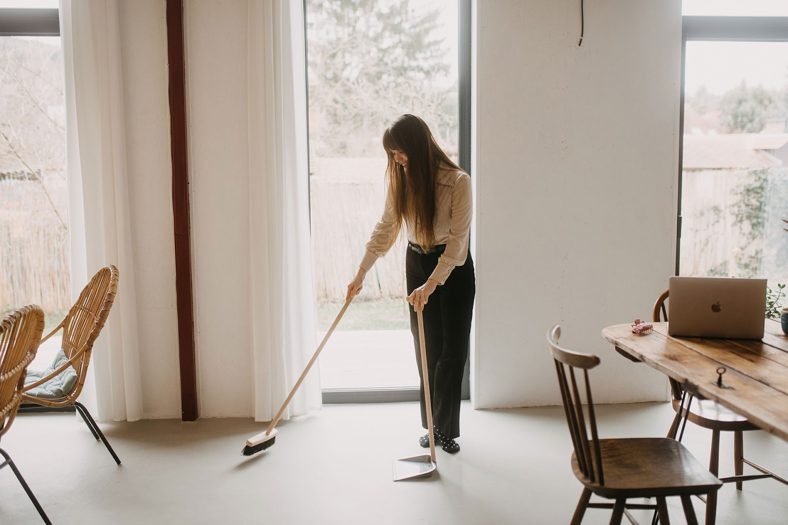 Woman sweeps floor in light and open home with floor to ceiling windows, timber table with laptop open,