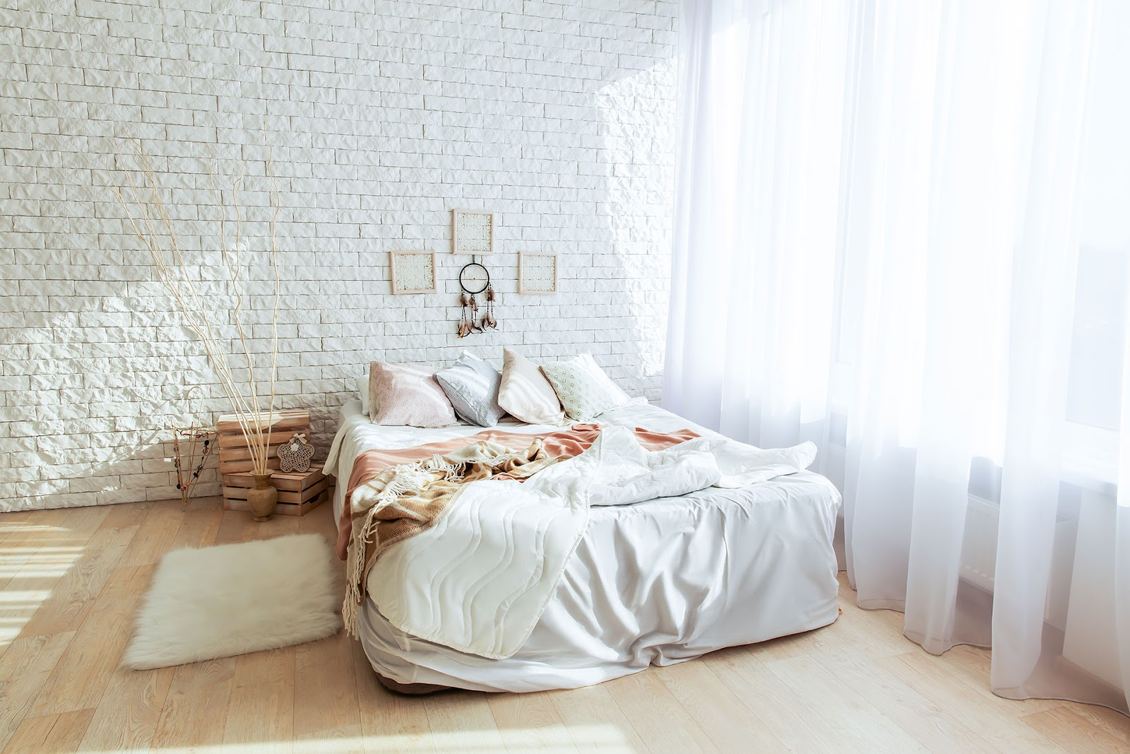 Boho style bedroom with sheer white curtains, exposed brick painted white, timber floorboards, white bed linen with light orange cushions and decorative timber crates beside the bed.