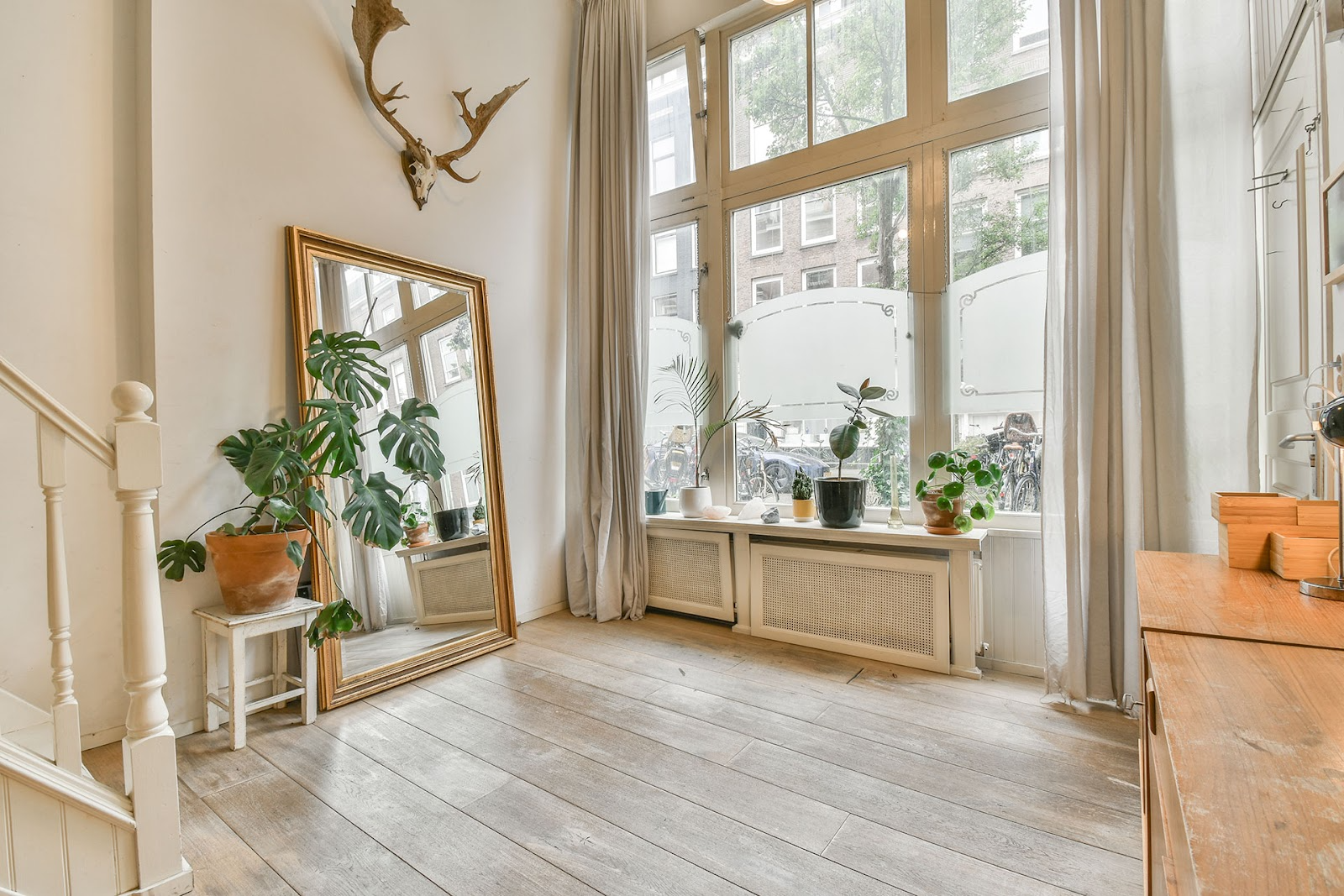 Eclectic landing space, greywashed timber floorboards, white and timber buffet and tables, large windows with curtains open, gold framed mirror leans agains wall with pot plant nearby.