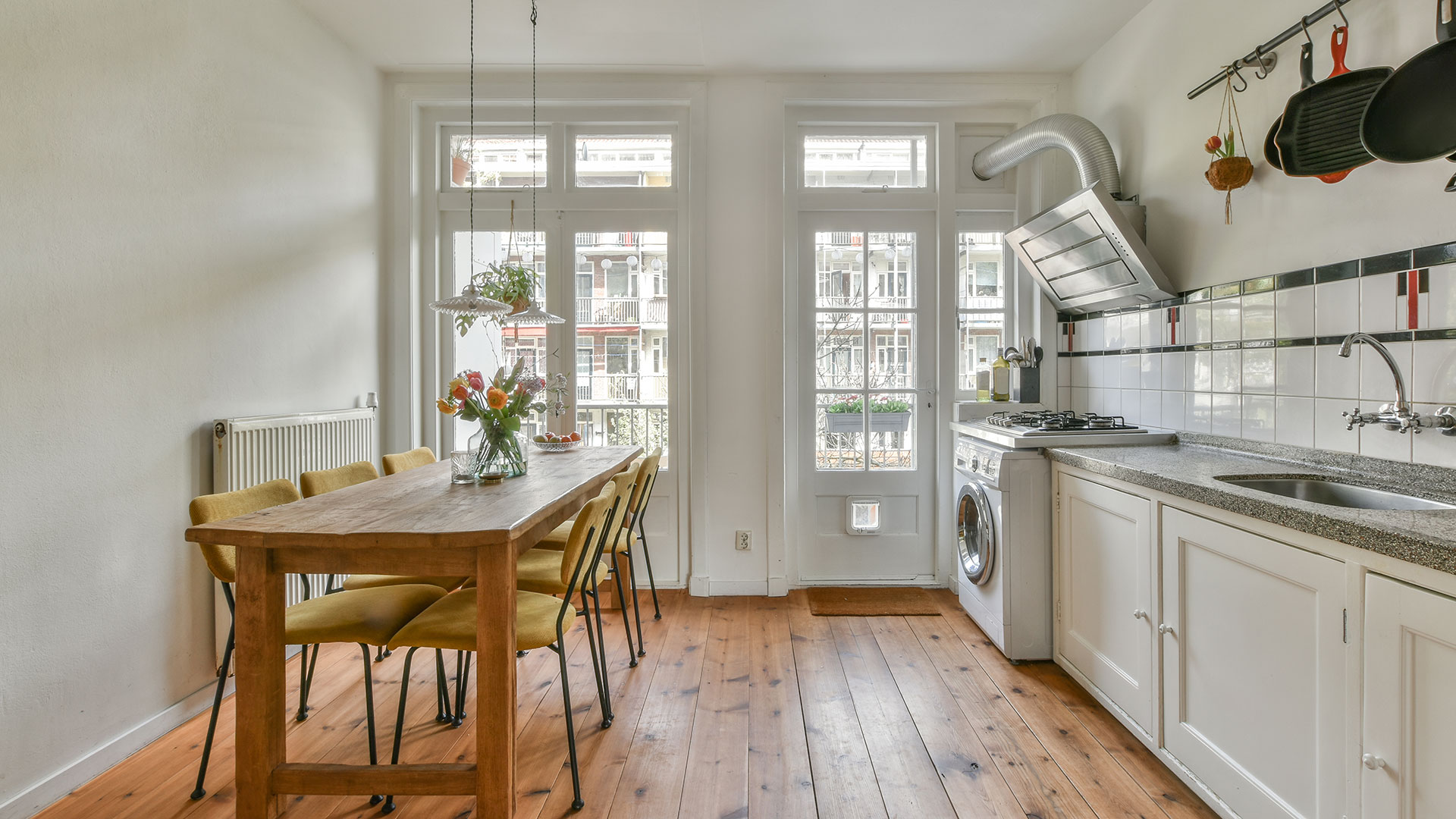 A modern apartment kitchen and dining area with mid-tone hardwood flooring bathed in natural light. The warm wood tones add depth and contrast to the sleek, contemporary design.