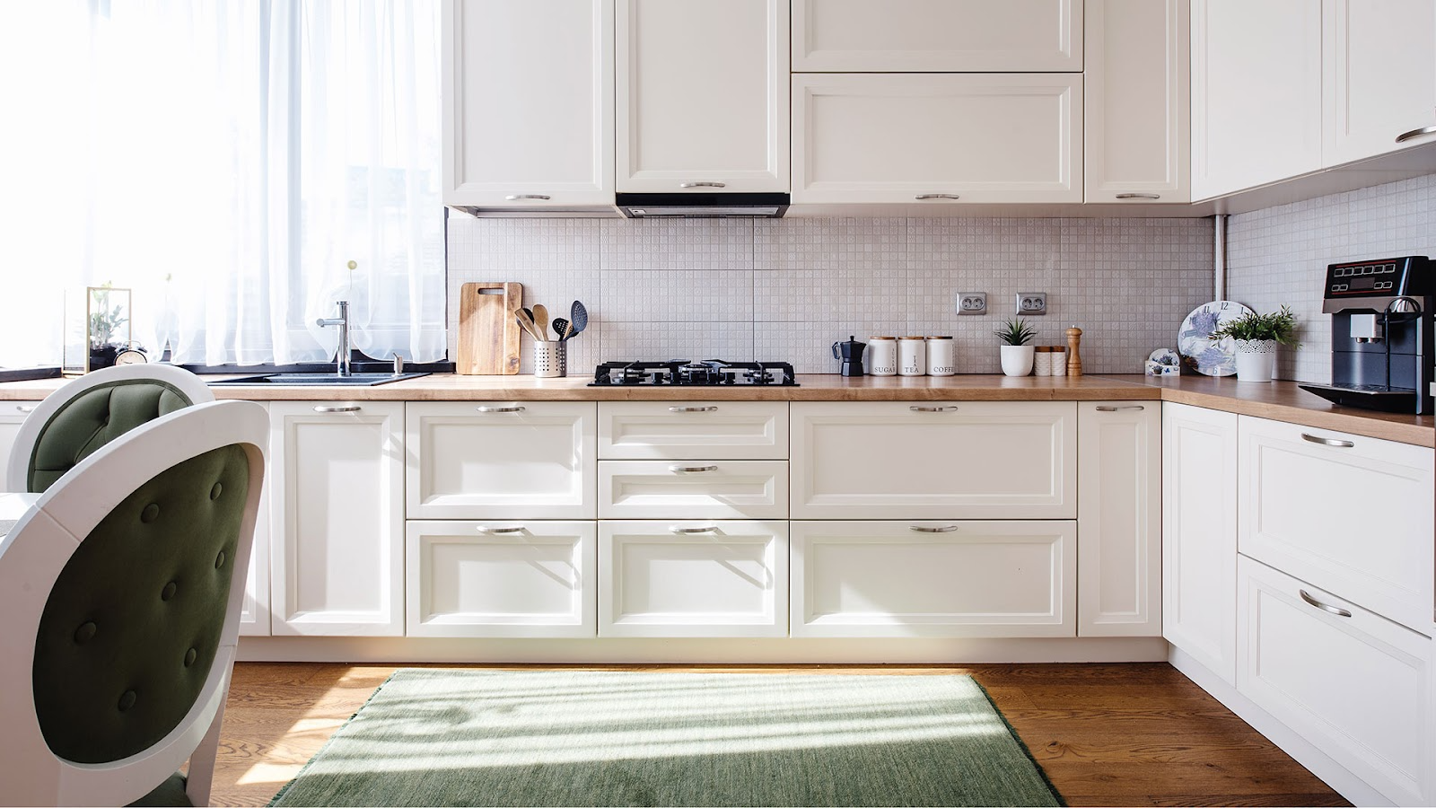 Kitchen with light timber benchtop and white cupboards, green and white dining chairs and green floor rug over timber floorboards.