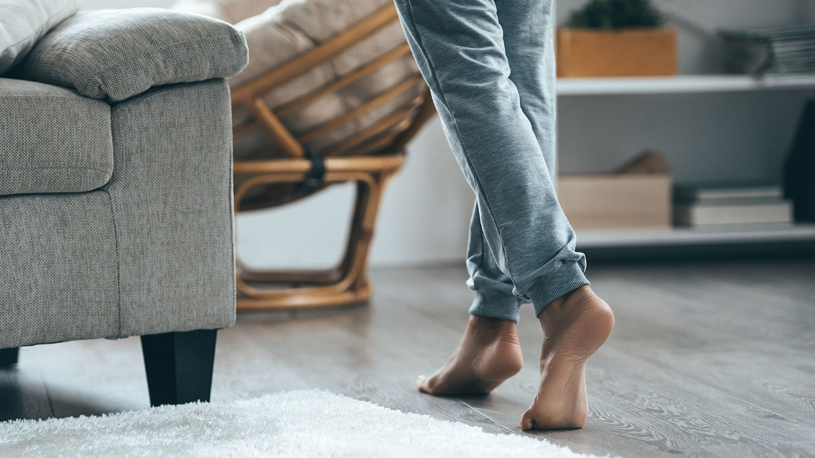 Woman walks barefoot through a lounge room with a grey couch, bookshelves, a white rug and dark floorboards.