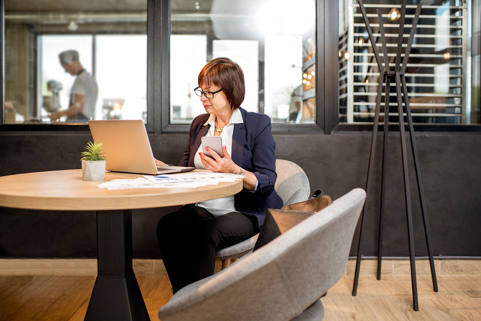 Business woman sits at round table with laptop and phone in hand.