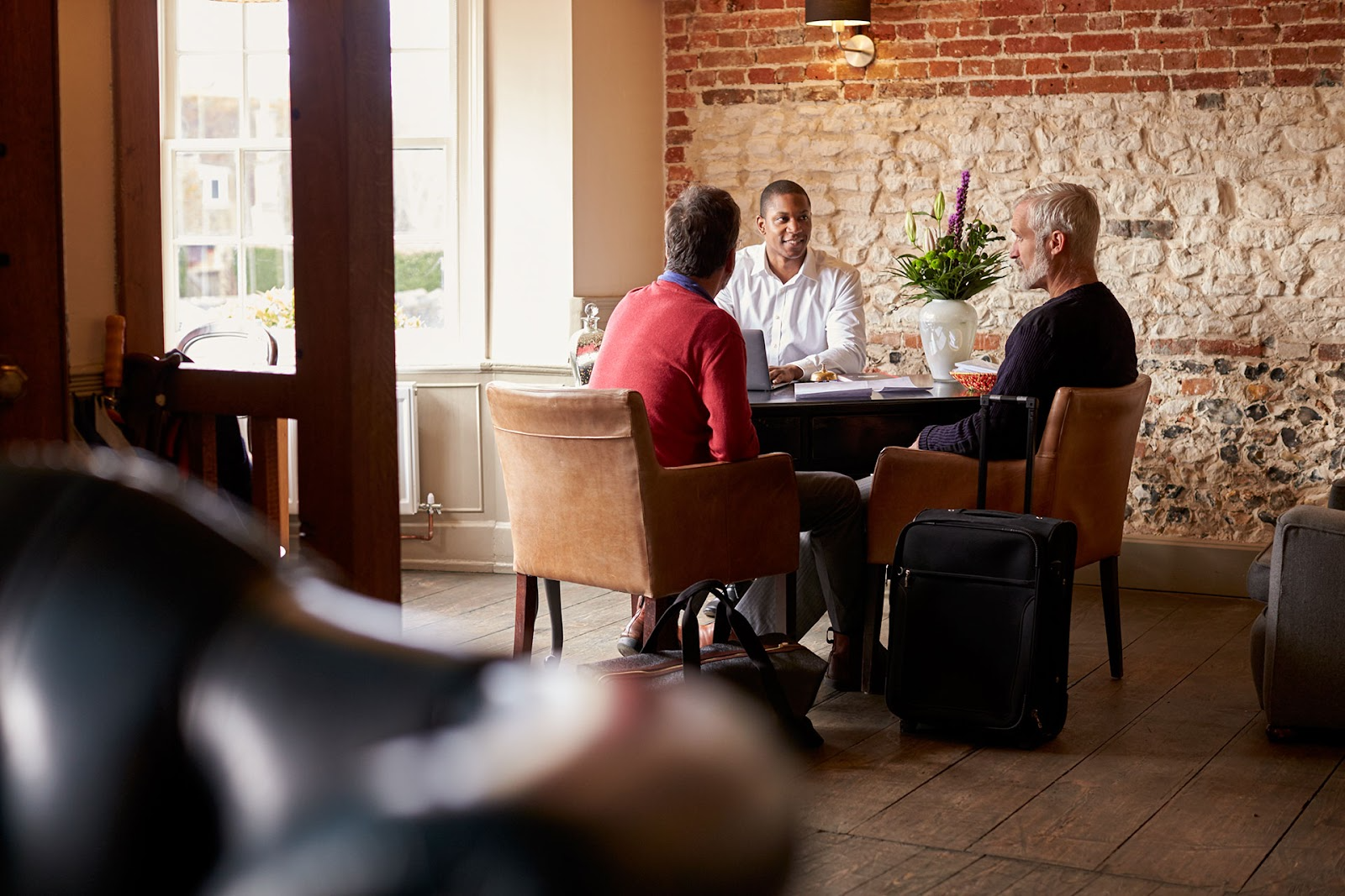 Three men sit around an informal hotel check-in desk. Exposed brick walls, hardwood timber floors.