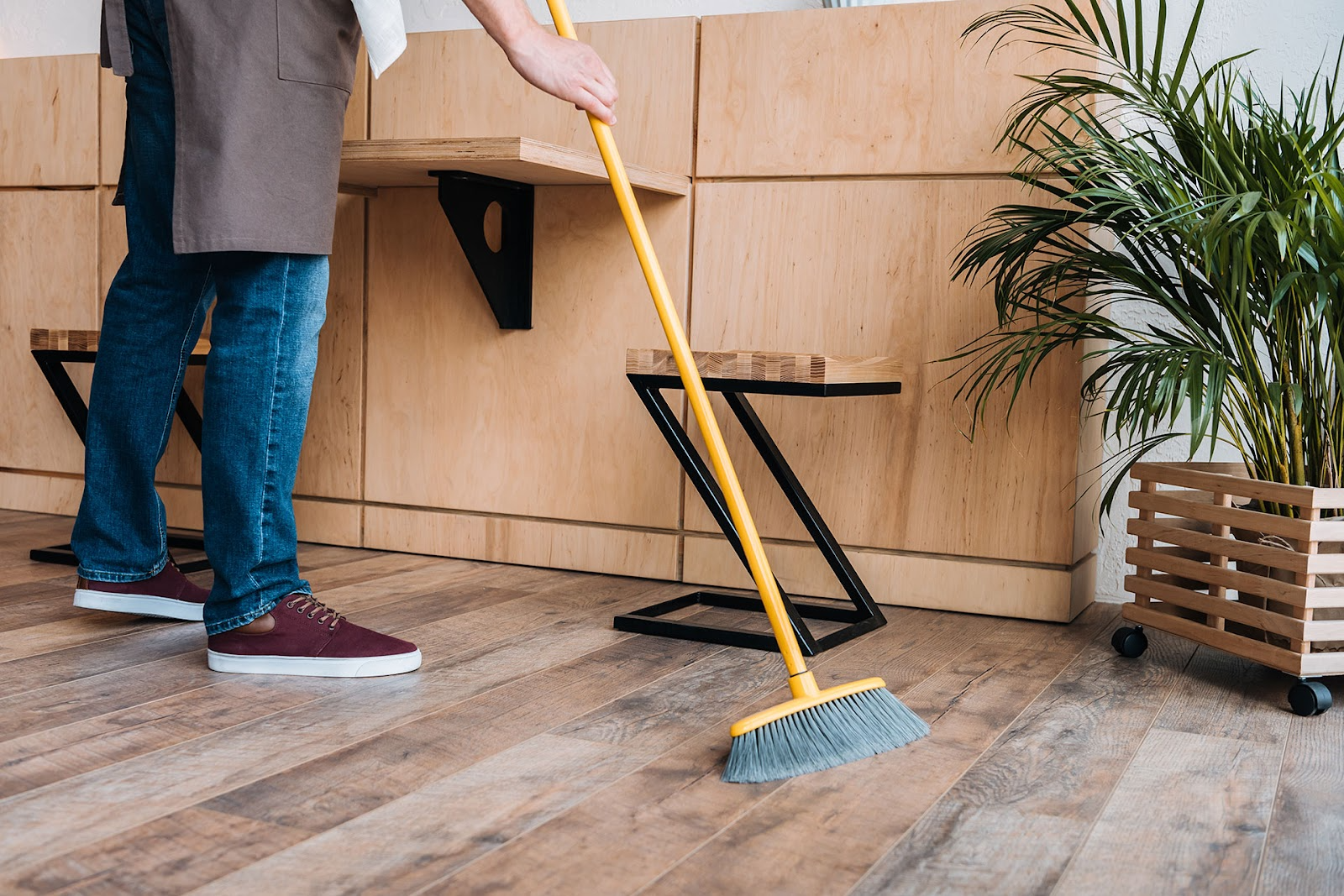 Young man sweeps the floor of a cafe. Timber wall with attached table and plant in corner.m