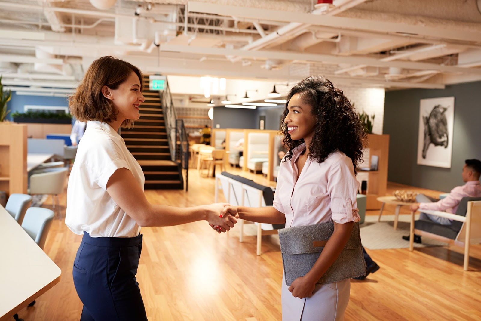 Two women shake ands in an open plan office building. Exposed pipes on ceiling, segmented booth seating, timber floors.