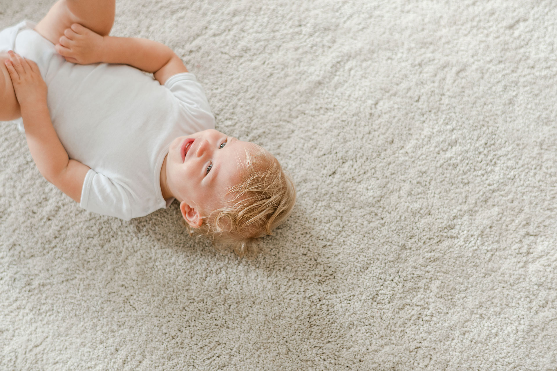 Smiling baby lays on soft, comfortable carpet flooring in a cozy home setting. Highlights warmth, safety, and comfort for family living.