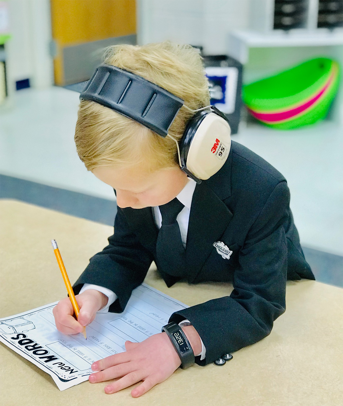 A young boy in a suit wearing noise cancelling headphones working on homework.