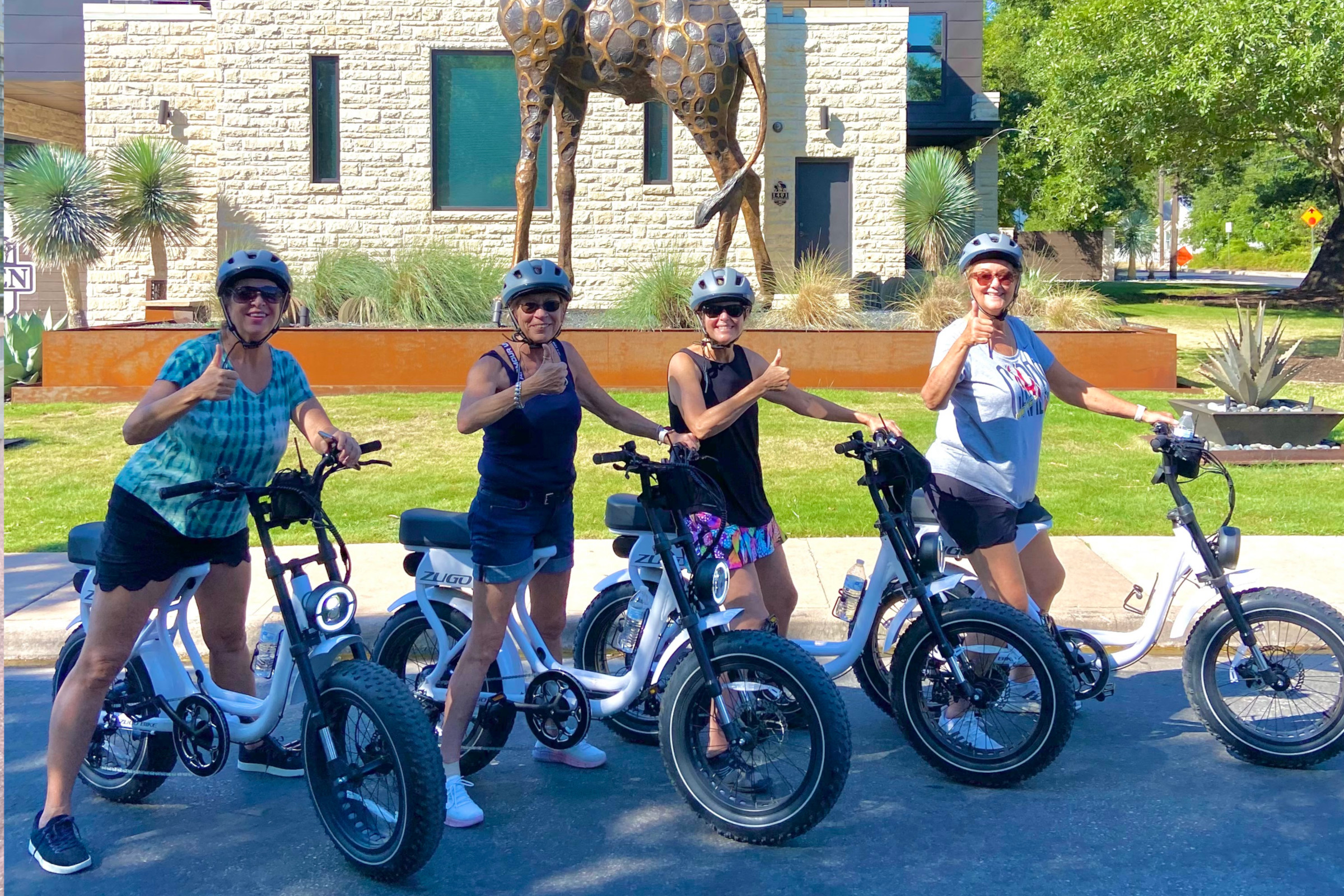 Four women wearing helmets on electric bikes giving thumbs up, posing on a sunny street with a building and large giraffe sculpture in the background.