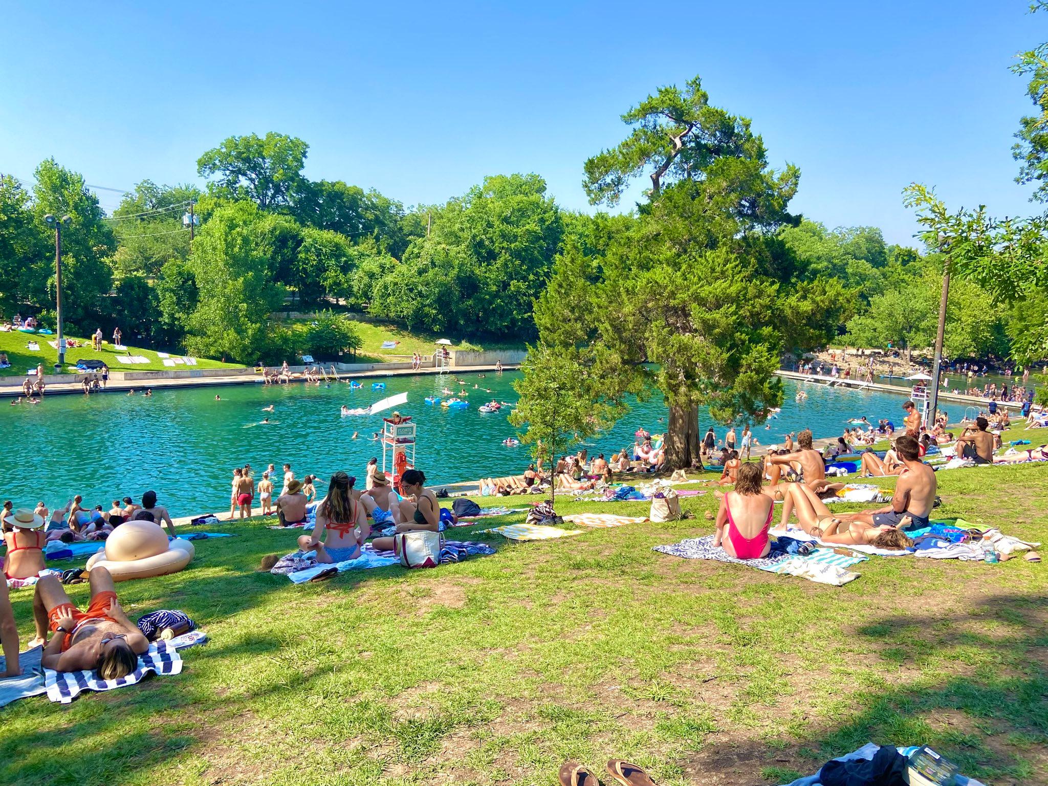 People relaxing on towels on grassy hill beside a natural swimming pool surrounded by trees on a sunny day.