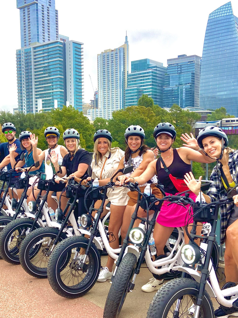Group of eight adults wearing helmets smiling and waving while standing with electric bikes on a city street with skyscrapers and trees in the background.